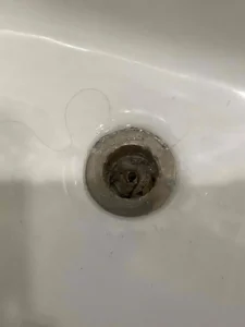 Close-up of a bathroom sink drain with dark buildup and a strand of hair nearby, showing how hair and grime collect and lead to clogs