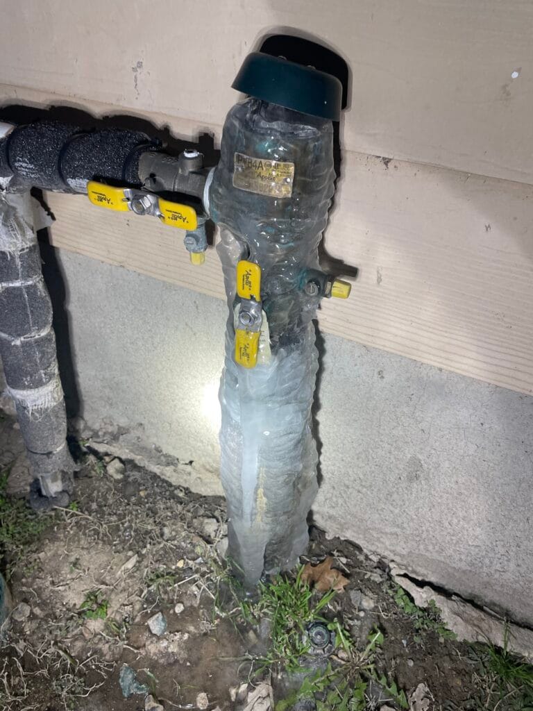 This photo shows a frozen outdoor backflow preventer on a plumbing system. The entire valve assembly and pipes are encased in a thick layer of ice, indicating extreme cold temperatures and a lack of proper insulation. The yellow-handled shutoff valves are visible and also frozen, while the black insulated pipe on the left appears to have better protection. The ground underneath is wet, suggesting that some ice may have already melted, possibly leading to a leak. The backflow preventer is labeled "Apollo", a common brand for such devices. The surrounding area includes a house's exterior wall and foundation, with a small access hole near the top. This is a clear example of freezing weather causing plumbing issues, which can result in pipe bursts and water damage if not addressed quickly. Insulating or wrapping this component in heat tape or a cover could have helped prevent freezing.