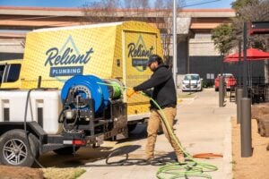 A Reliant Plumbing technician prepares a high-pressure hose from a hydro jetting machine mounted on a trailer, parked beside a yellow Reliant Plumbing van. The scene takes place in a commercial parking lot under a clear blue sky.