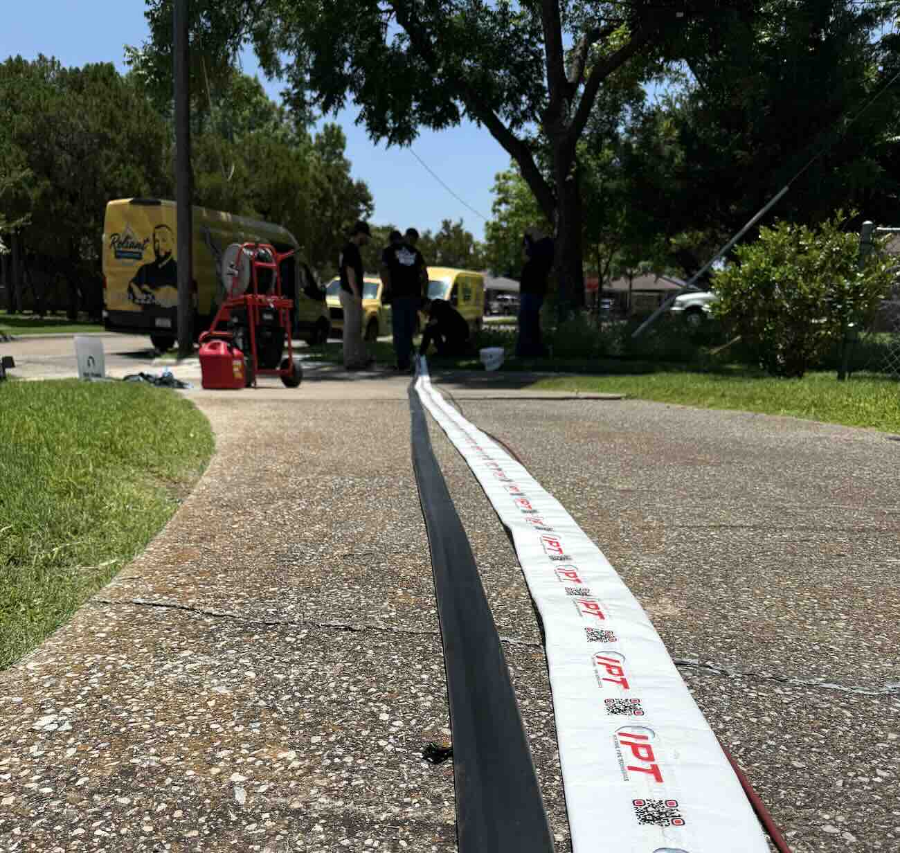 Trenchless pipe lining equipment laid out on a residential driveway, with Reliant Plumbing technicians preparing for a no-dig sewer line repair. Yellow service vans and plumbing tools are visible in the background under a sunny sky.