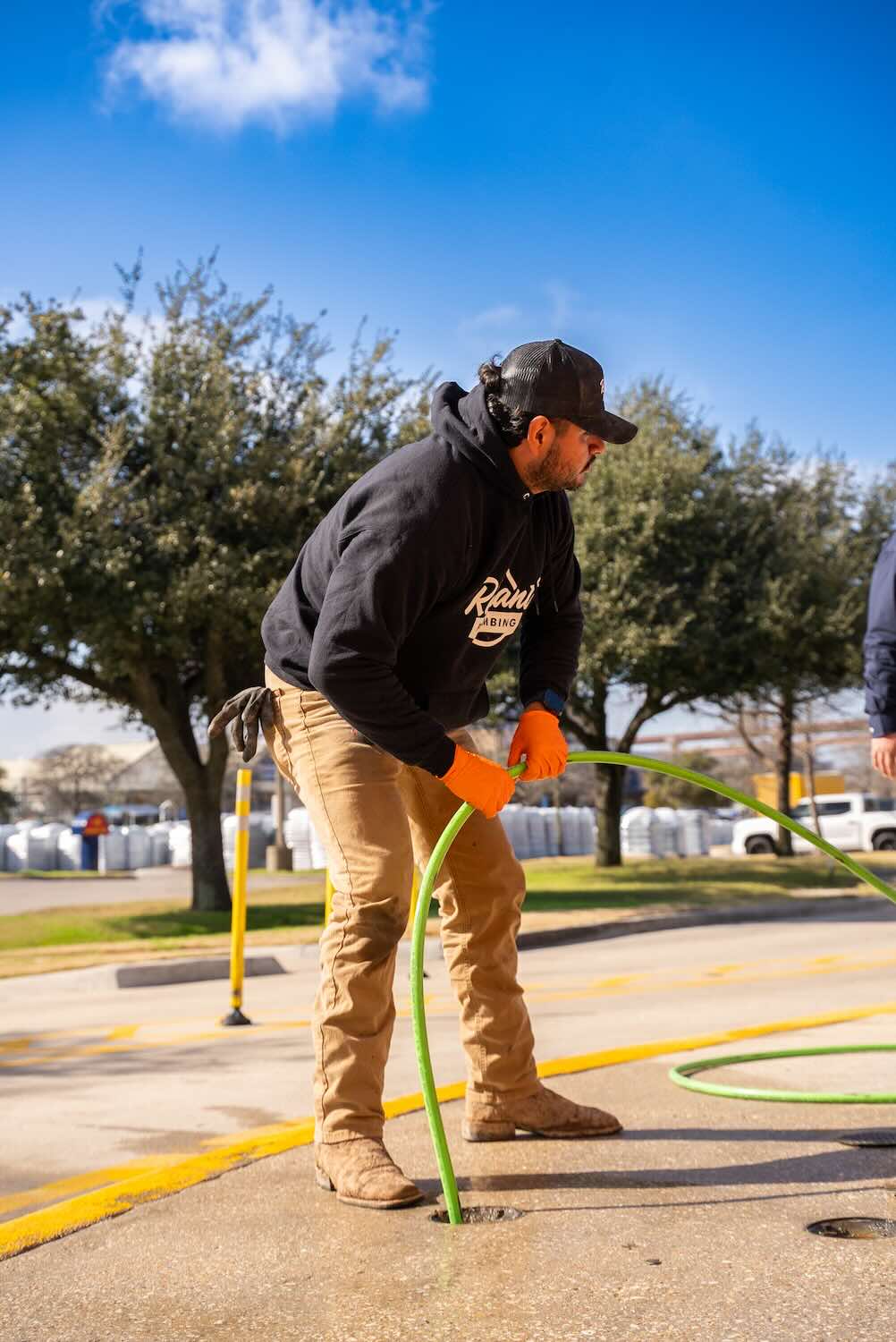 A Reliant Plumbing technician inserts a green hydro jetting hose into a manhole or outdoor sewer cleanout. The worker is wearing a black hoodie, orange gloves, and muddy boots, performing the service on a sunny day with trees and parked vehicles in the background.