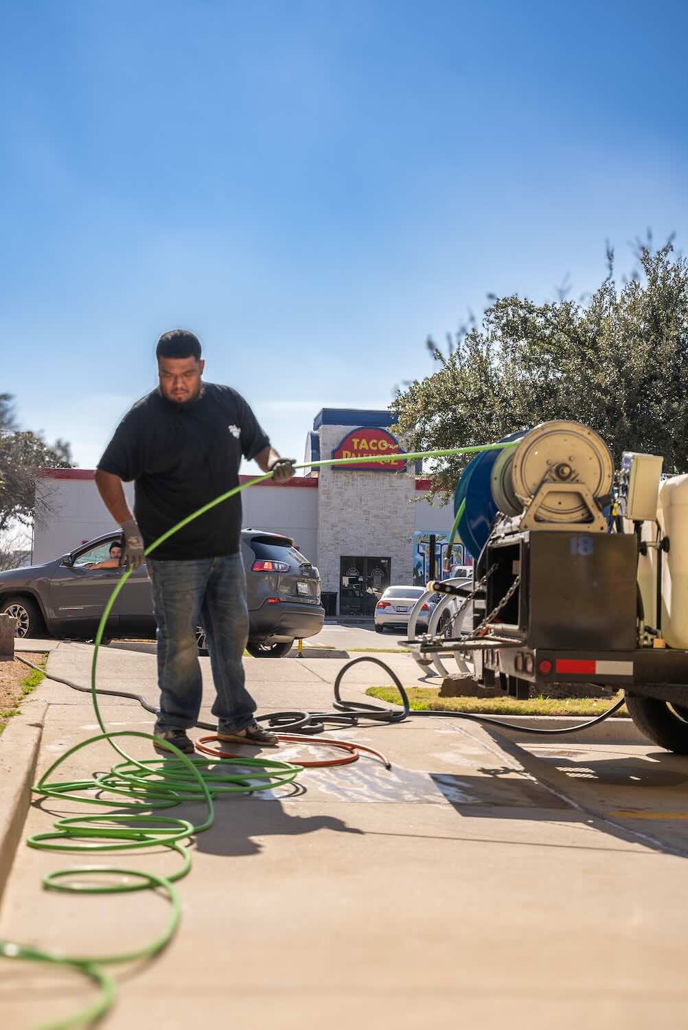 sewer-line-cleaning A Reliant Plumbing technician prepares a green hydro jetting hose beside a commercial trailer-mounted jetter unit on a sunny day. In the background, cars and a Taco Palenque restaurant are visible, indicating a busy commercial area.