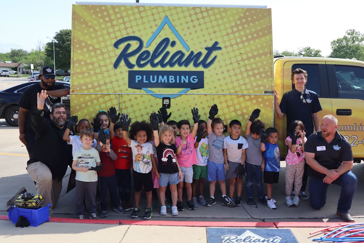 Group of young children standing in front of a Reliant Plumbing truck with several Reliant team members, all smiling and waving during a community or school career day event.
