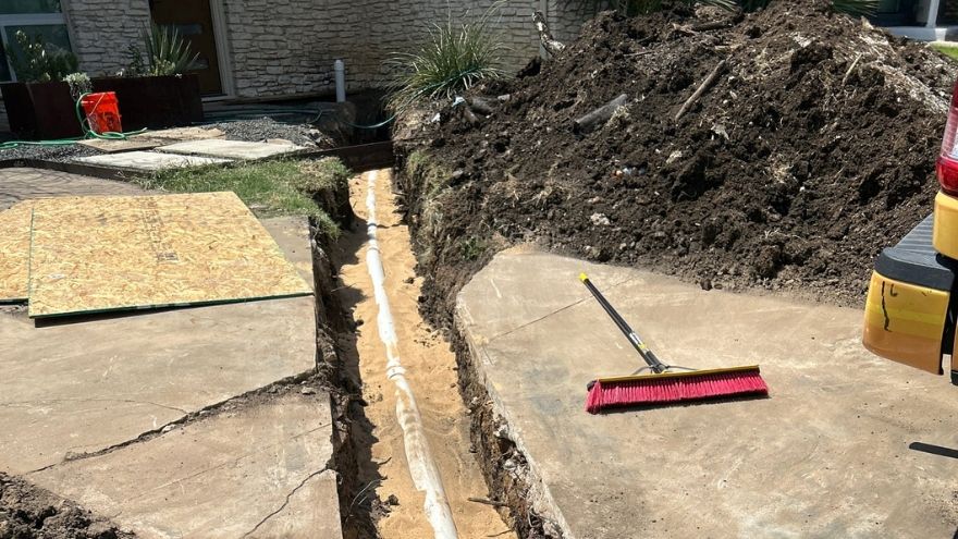 driveway dug up with a new pvc pipe laid in the trench with a broom on the right of the trench, a large pile of dirt on the upper right side, and plywood on the left