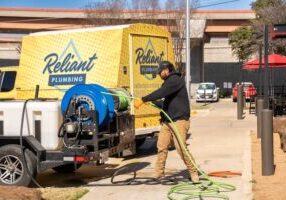 A Reliant Plumbing technician prepares a high-pressure hose from a hydro jetting machine mounted on a trailer, parked beside a yellow Reliant Plumbing van. The scene takes place in a commercial parking lot under a clear blue sky.